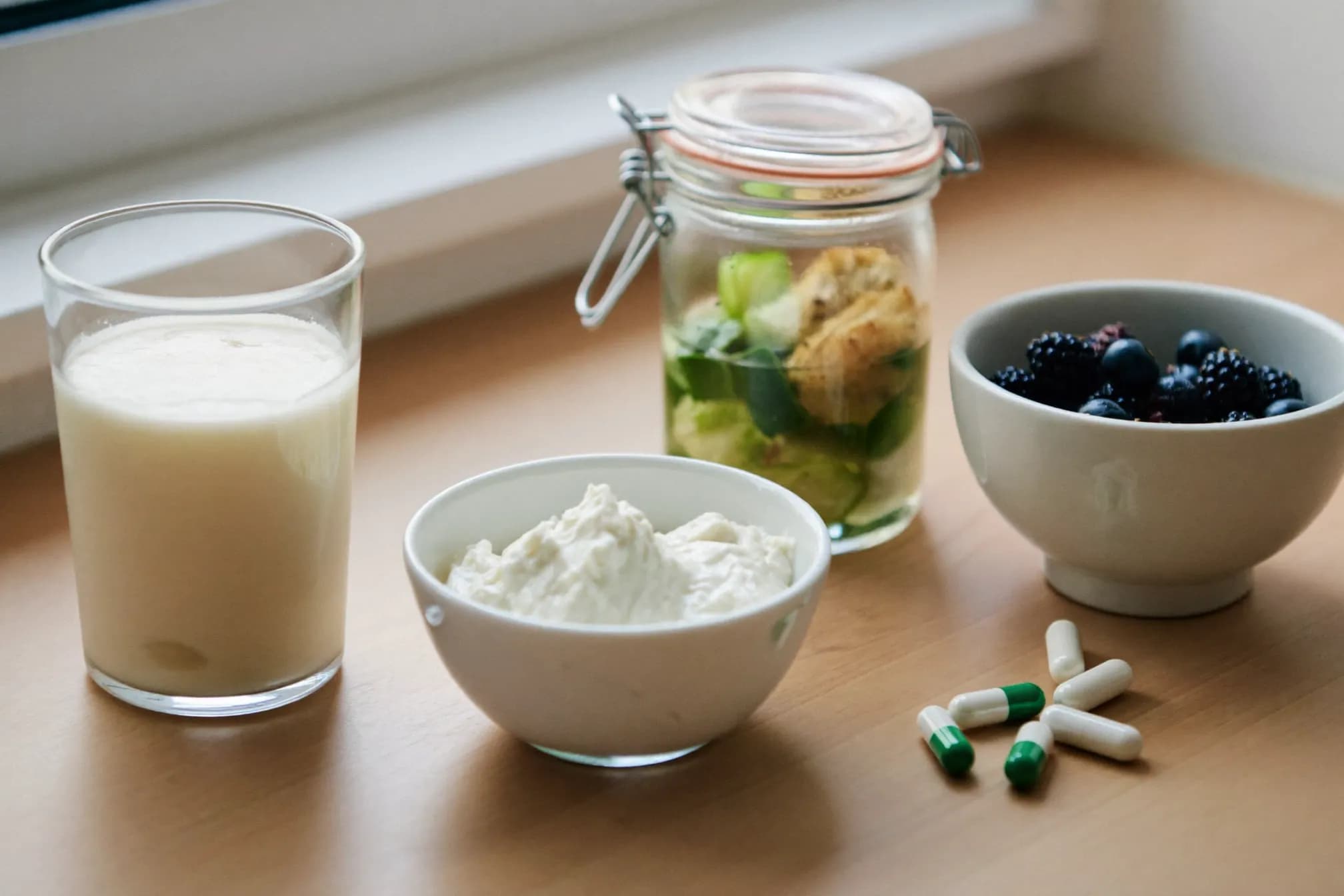 A glass of kefir and a bowl of natural yogurt with fresh berries beside probiotic capsules on a light kitchen counter.