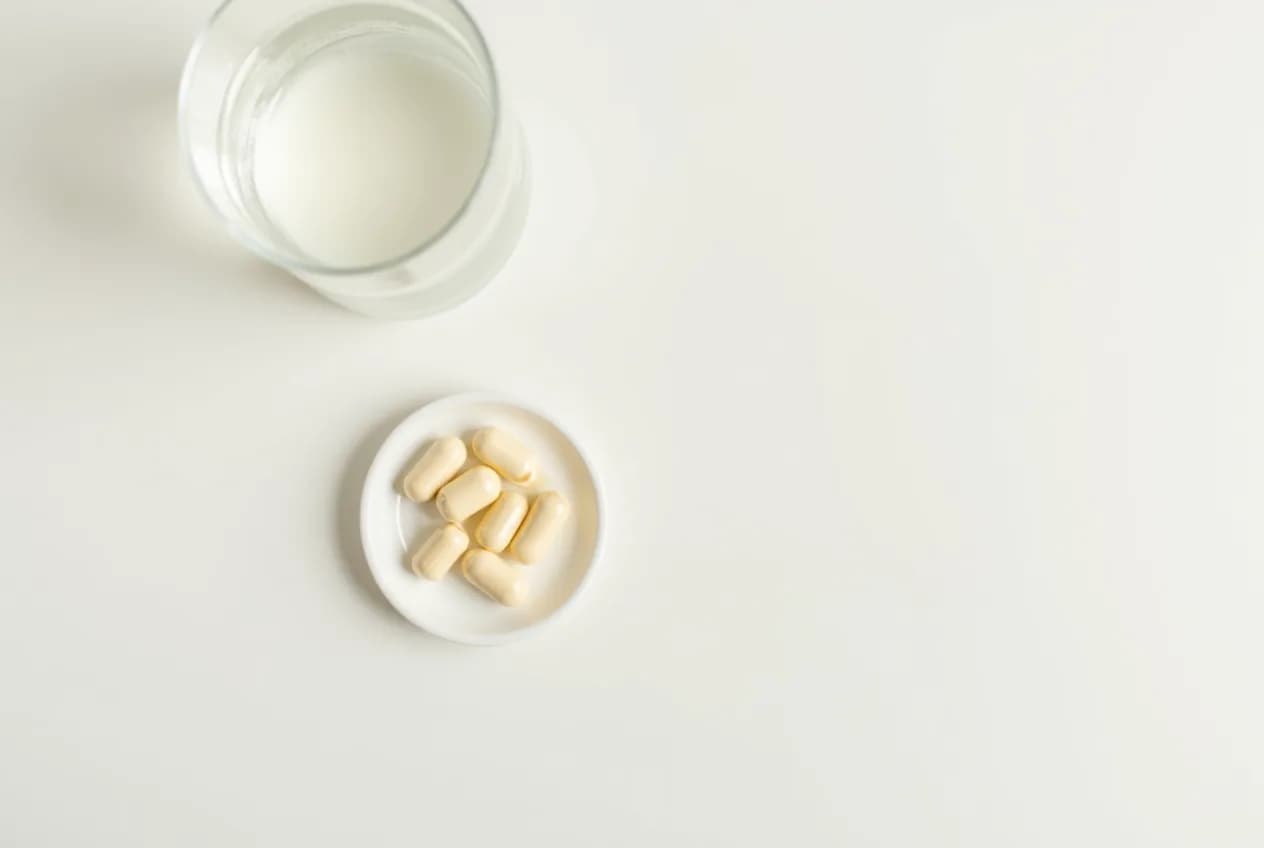 An overhead flat-lay of pale supplement capsules in a small dish beside a glass of water