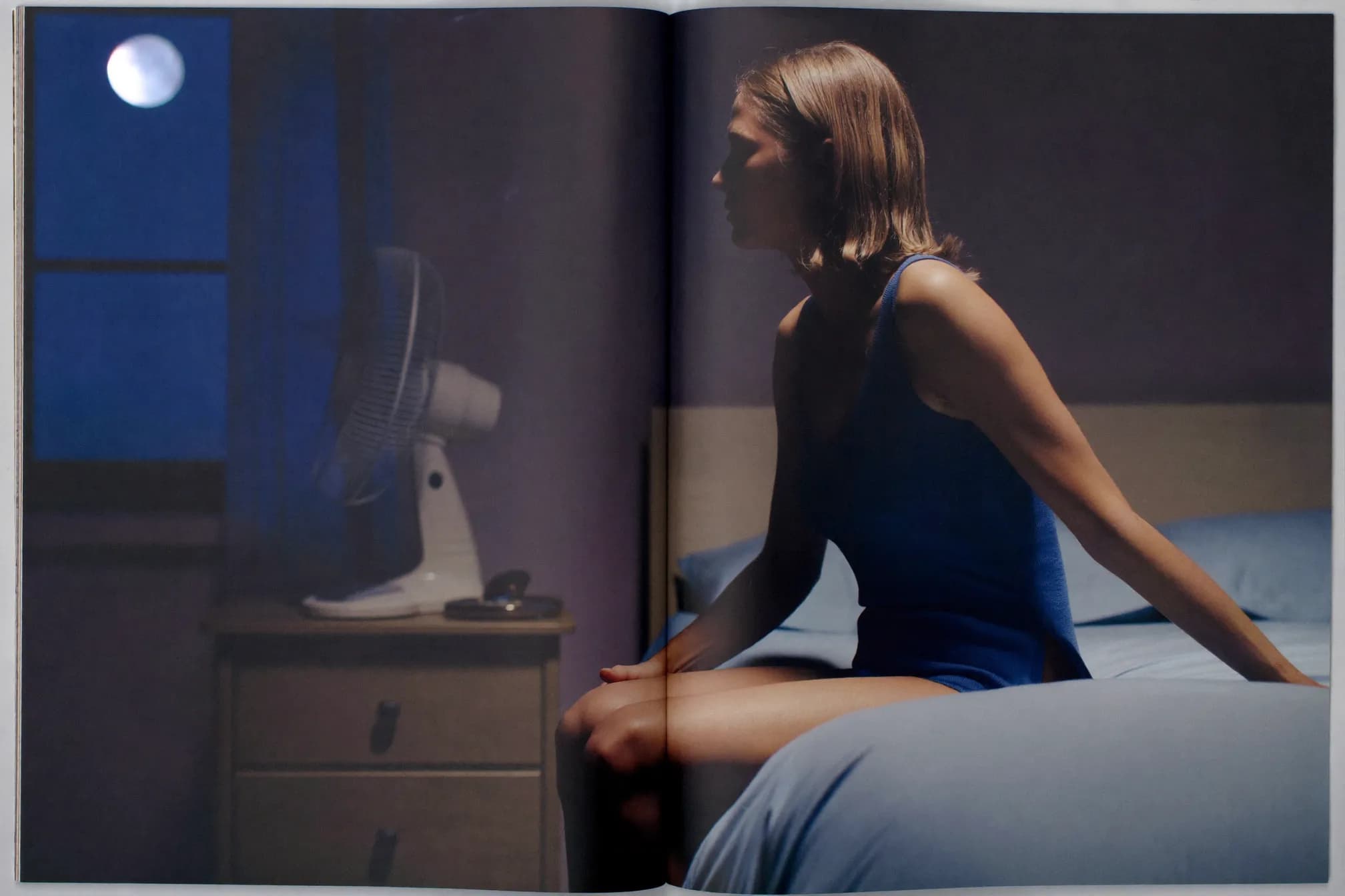 A woman in her late forties sitting on the edge of her bed at night by cool moonlight.