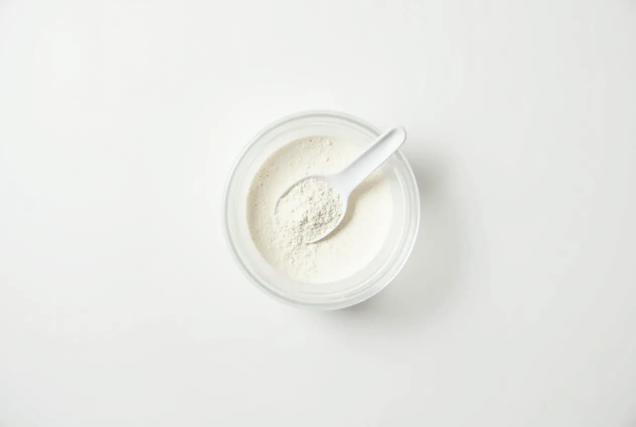 An overhead flat-lay of a scoop of white creatine powder, a tub, and a glass of water on a neutral surface
